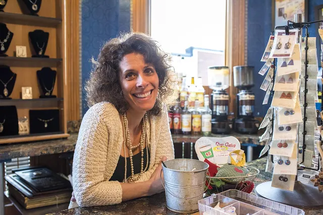 Smiling boutique owner with curly hair leans on a shop counter beside an earring display rack and necklace shelves