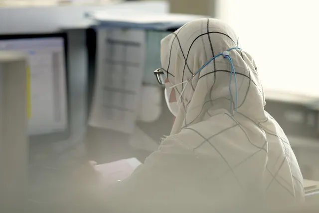 Woman in a plaid hijab, glasses and face mask reviewing papers at an office desk beside a computer monitor
