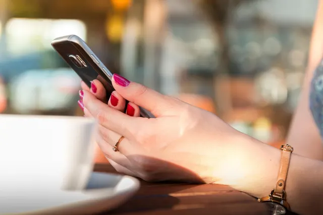 Woman with red nail polish holding a smartphone at a sunny café table beside a white coffee cup