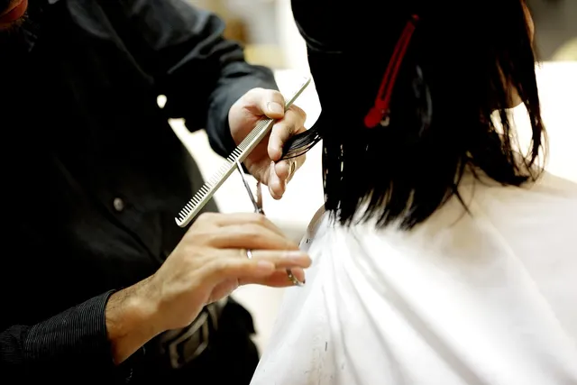 Stylist in black shirt trimming a client's wet dark hair with scissors and comb, client draped in a white salon cape