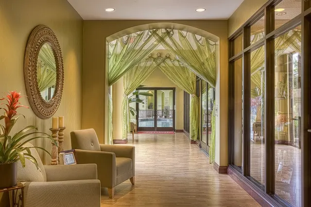 Hotel hallway with beige armchairs, ornate round mirror, red bromeliad, green draped archway and glass doors to a pool