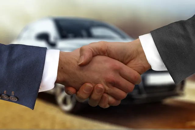 Two men in suit jackets shaking hands in front of a blurred silver car, suggesting a closed car dealership sale