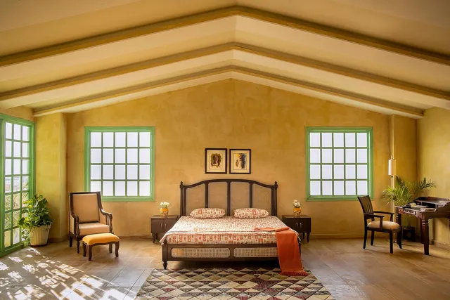 Sunlit bedroom with a cane-panel wooden bed, armchair and ottoman, writing desk, and patterned rug under green-framed windows