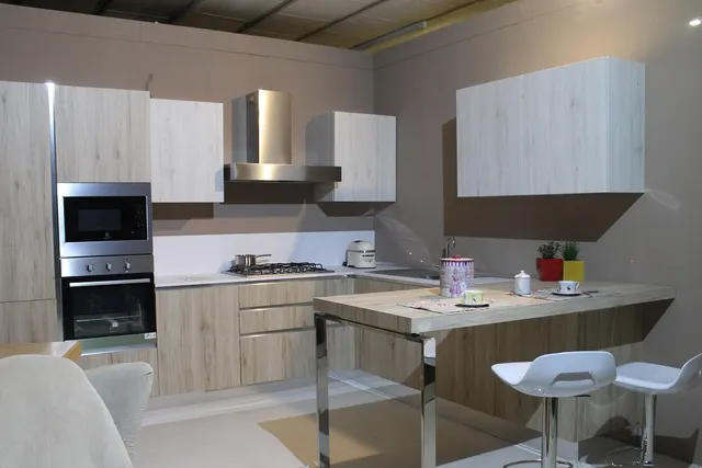Showroom kitchen display with light wood cabinets, stainless range hood, built-in oven and breakfast bar with white stools
