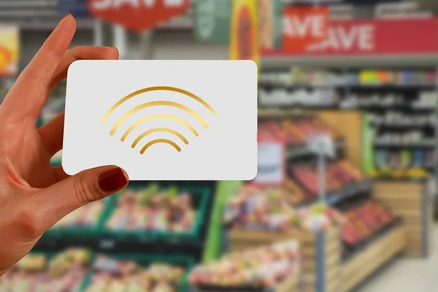 Hand holding a white card with a gold contactless payment symbol in front of blurred grocery store produce aisles
