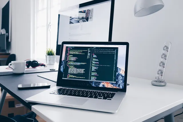 Laptop showing code on a white desk with a large monitor, smartphone, coffee mug, headphones and small plant