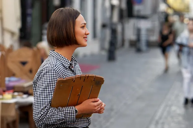 Smiling restaurant hostess in a gingham shirt holds a wooden menu on a cobblestone street beside outdoor café tables