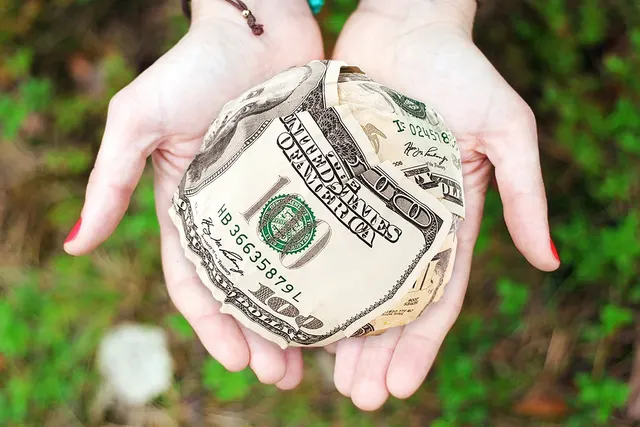 Two open palms holding a ball of crumpled US $100 bills over a blurred background of green grass and leaves