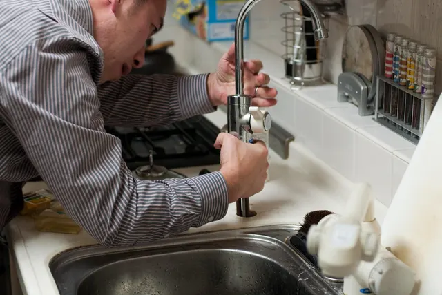 Man in a striped shirt repairing a chrome kitchen faucet over a stainless steel sink in a tiled home kitchen