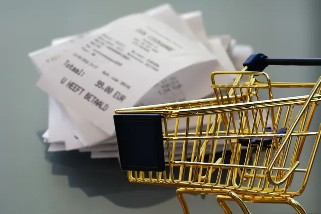 Miniature gold shopping cart in front of a pile of printed receipts, one showing a 99.00 EUR total