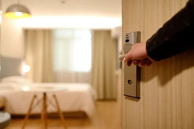 Hand opening a hotel room door with a keycard lock, revealing a softly lit guest room with a made bed and curtains