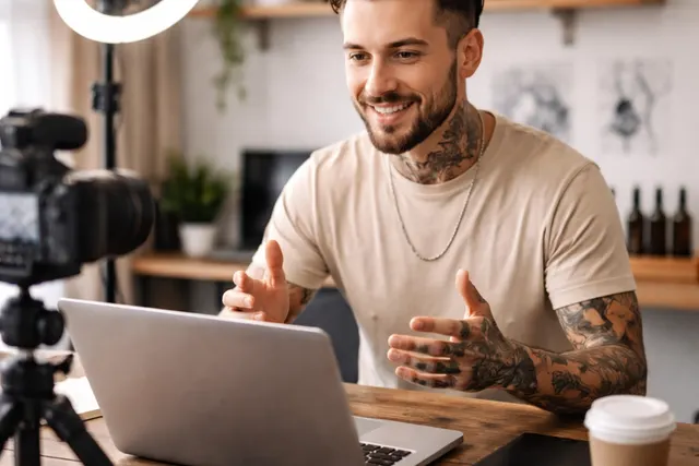 Smiling tattooed creator gestures at a laptop on a wooden desk, filmed by a camera with a ring light in a home studio