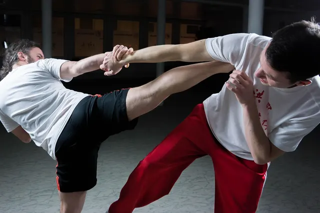 Two men sparring in a martial arts studio, one blocking a wrist grab while landing a side kick to the other's midsection
