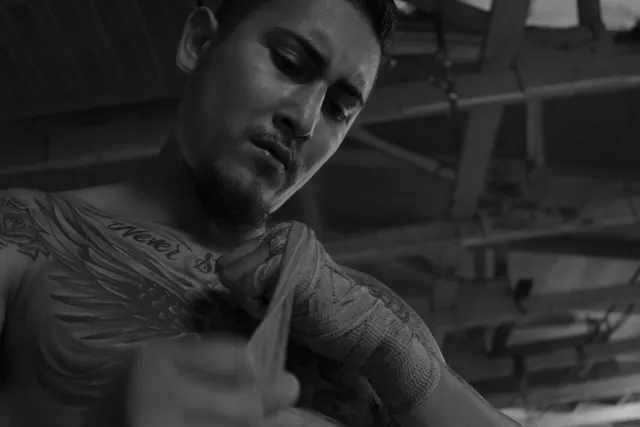 Black-and-white photo of a shirtless, tattooed boxer wrapping his hands under the wooden roof beams of a gym