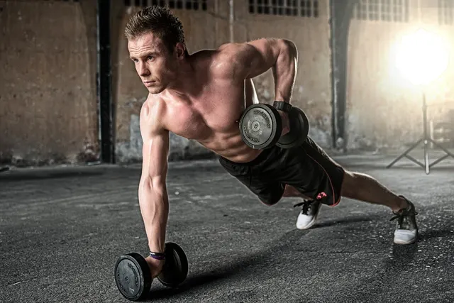 Shirtless man doing a one-arm dumbbell row in plank position on concrete floor of an industrial gym space