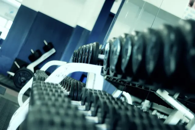 Close-up of rows of black hex dumbbells on a white rack in a gym, with weight plates and blue walls in the background