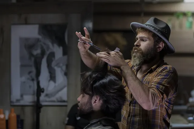 Bearded stylist in a fedora and plaid shirt trims a client's dark hair with scissors and comb in a rustic salon