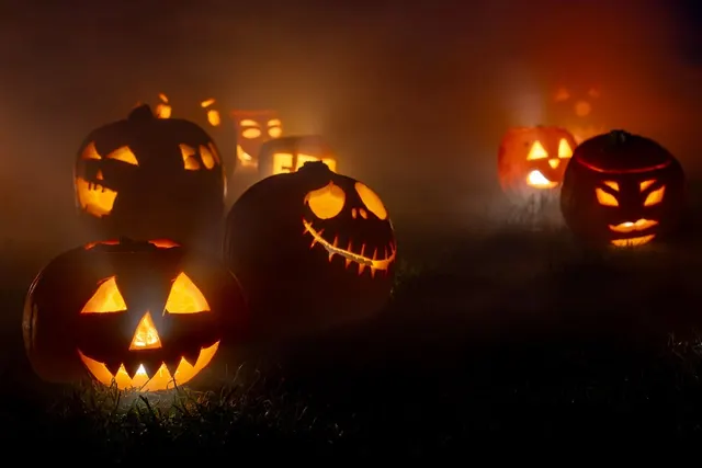 Glowing carved jack-o'-lanterns with spooky faces sitting on grass in a foggy field at night