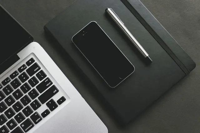 Overhead view of a silver laptop keyboard beside a smartphone and metal pen on a black notebook on a dark desk