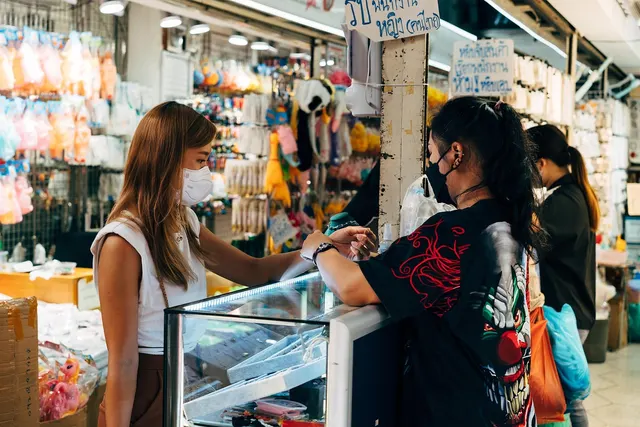Masked shopper in a white top at a glass display counter as a woman in black hands over an item in a busy market stall