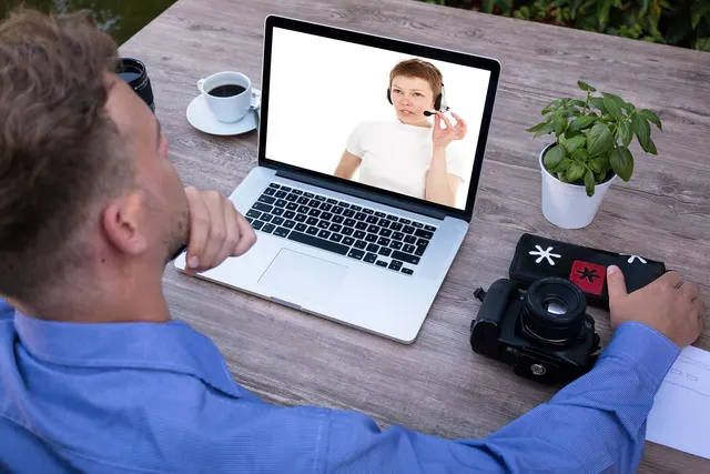 Man in blue shirt on a video call with a woman wearing a headset on a laptop at a wooden desk with coffee and a camera