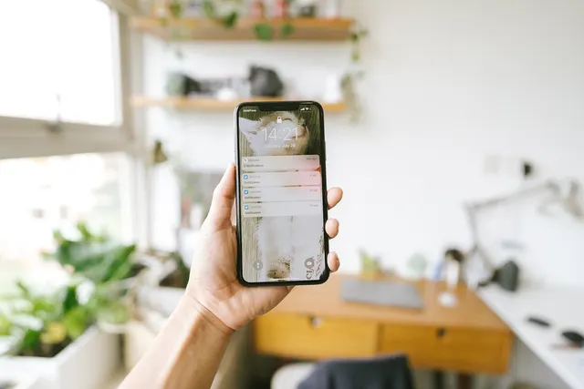 Hand holding a smartphone showing lock screen notifications in a bright home office with plants and a wooden desk