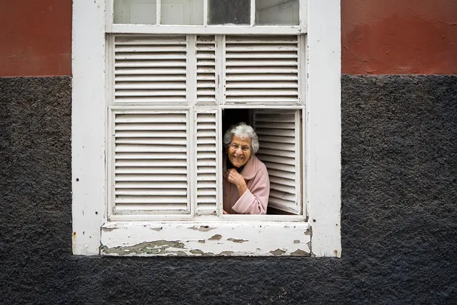 Smiling elderly woman with glasses in a pink coat peeking out of a white shuttered window on a weathered wall