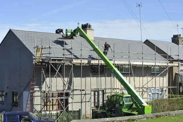 Roofers working on a slate roof of a semi-detached house surrounded by scaffolding, with a green telehandler lifting materials