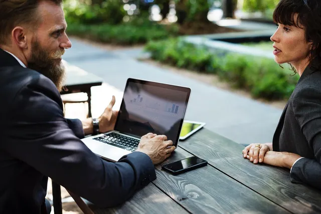 Bearded man in a suit shows charts on a laptop to a woman at an outdoor wooden table with a phone and tablet