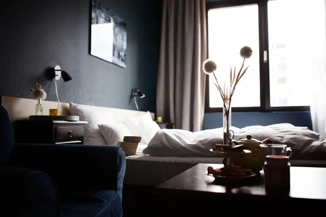 Dimly lit hotel room with a made bed, wall lamps, armchair and a table set with a teapot and pastries by the window