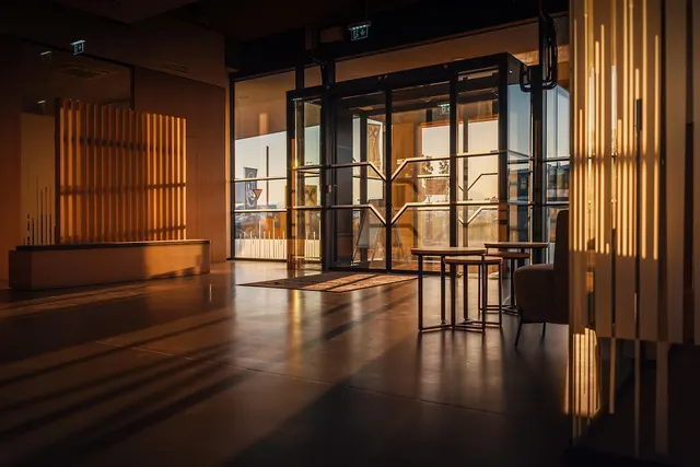 Empty hotel lobby at sunset with glass entrance doors, wooden slat walls, small tables and warm light on the floor