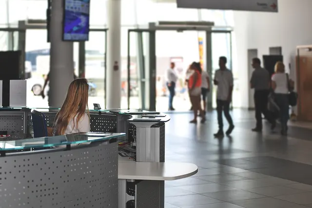 Receptionist with long hair seated at a glass-topped front desk in a bright lobby as guests walk past