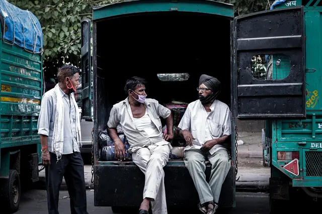 Three men in face masks talk while two sit on the open tailgate of a green cargo truck parked between other trucks