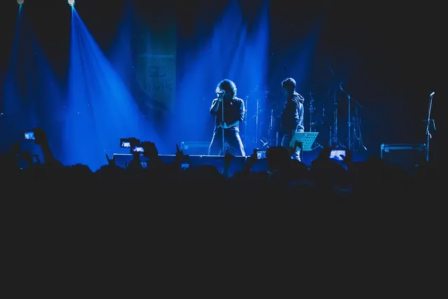 Singer performs on a blue-lit concert stage as silhouetted fans in the crowd hold up phones to record the show