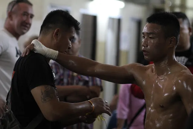 Sweaty shirtless fighter with taped hand rests his arm on a cornerman's shoulder backstage at a fight event