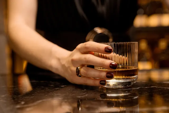 Hand with dark red manicured nails holding a ribbed glass of whiskey on a polished marble bar counter