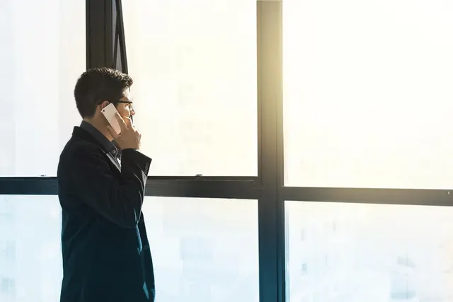 Businessman in a dark suit and glasses talking on a smartphone beside large sunlit office windows