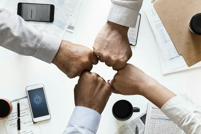 Four business colleagues bump fists over a white desk with smartphones, financial reports, notes and coffee cups
