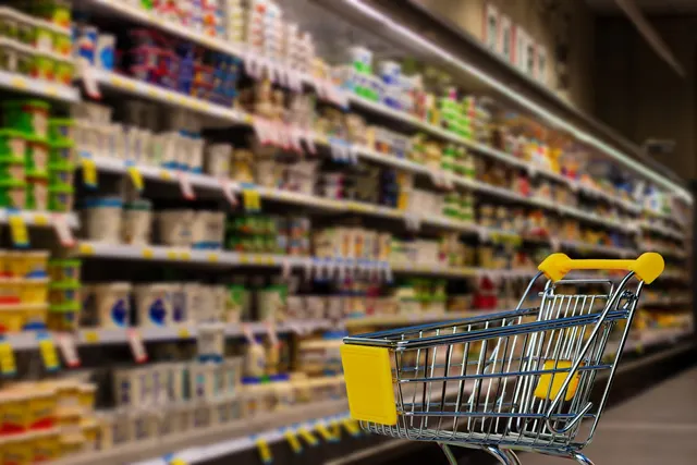 Empty shopping cart with yellow handle in a grocery store aisle beside blurred refrigerated shelves of dairy products