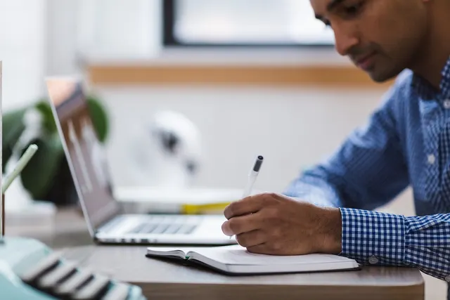 Man in a blue checkered shirt writing in a notebook at a desk beside an open laptop in a bright office