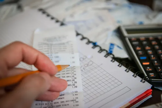 Hand holding a pencil over a printed receipt on a grid notebook beside a calculator and scattered receipts