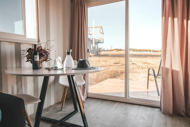 Guest cabin interior with a round table set with wine, glasses and a carafe beside a sliding door to a sunny desert view