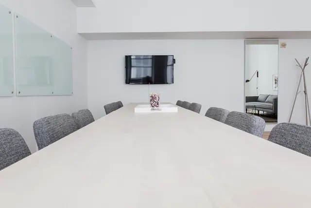 Empty bright conference room with a long white table, gray upholstered chairs, wall-mounted TV and glass whiteboards