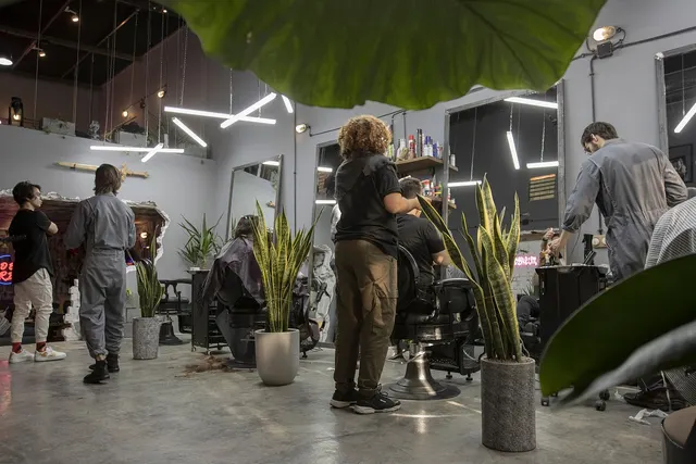 Barbers in gray coveralls cutting clients' hair at chairs in a modern barbershop with mirrors, LED lights and potted plants