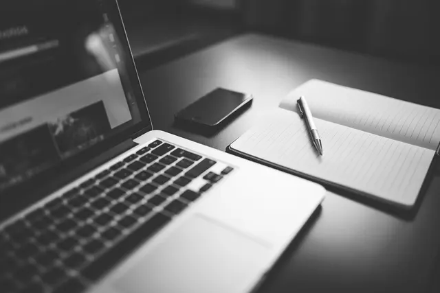 Black-and-white desk with an open laptop, smartphone, and blank lined notebook with a pen, ready for comparing quotes