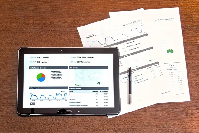 Tablet showing an analytics dashboard with charts beside printed performance reports and a pen on a wooden desk