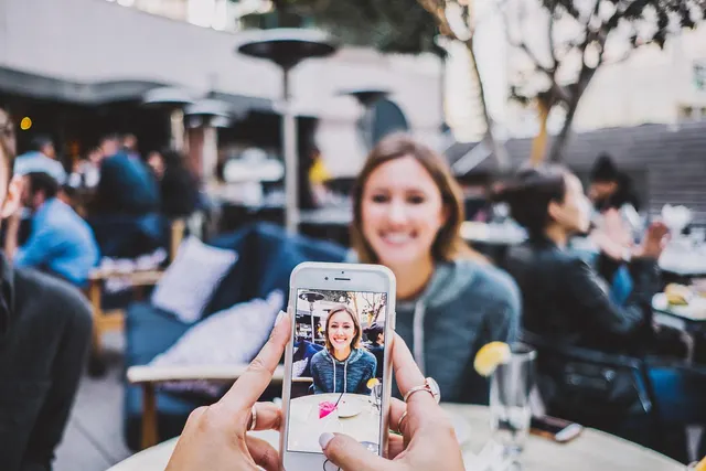 Hands holding a smartphone to photograph a smiling woman at a table on a busy outdoor restaurant patio