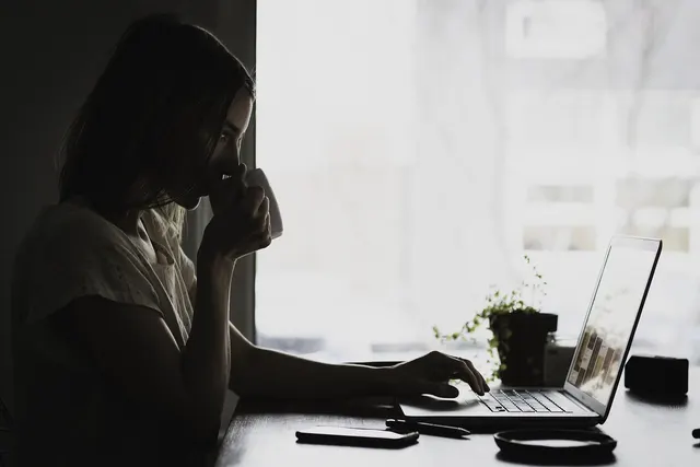 Woman sipping from a mug while typing on a laptop at a desk by a bright window, with a phone and small plant nearby