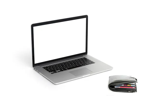 Open silver laptop with blank white screen beside a black leather wallet stuffed with payment cards on white