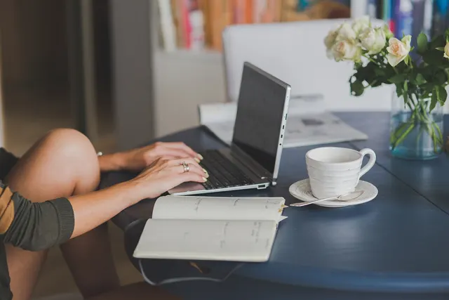 Woman typing on a laptop at a blue table with an open notebook, white coffee cup and vase of cream roses